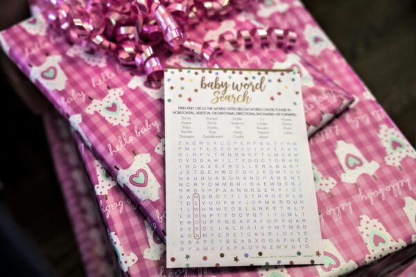 Baby word search game card placed on pink gift wrapping with a heart-patterned onesie design and pink ribbon at a baby shower.