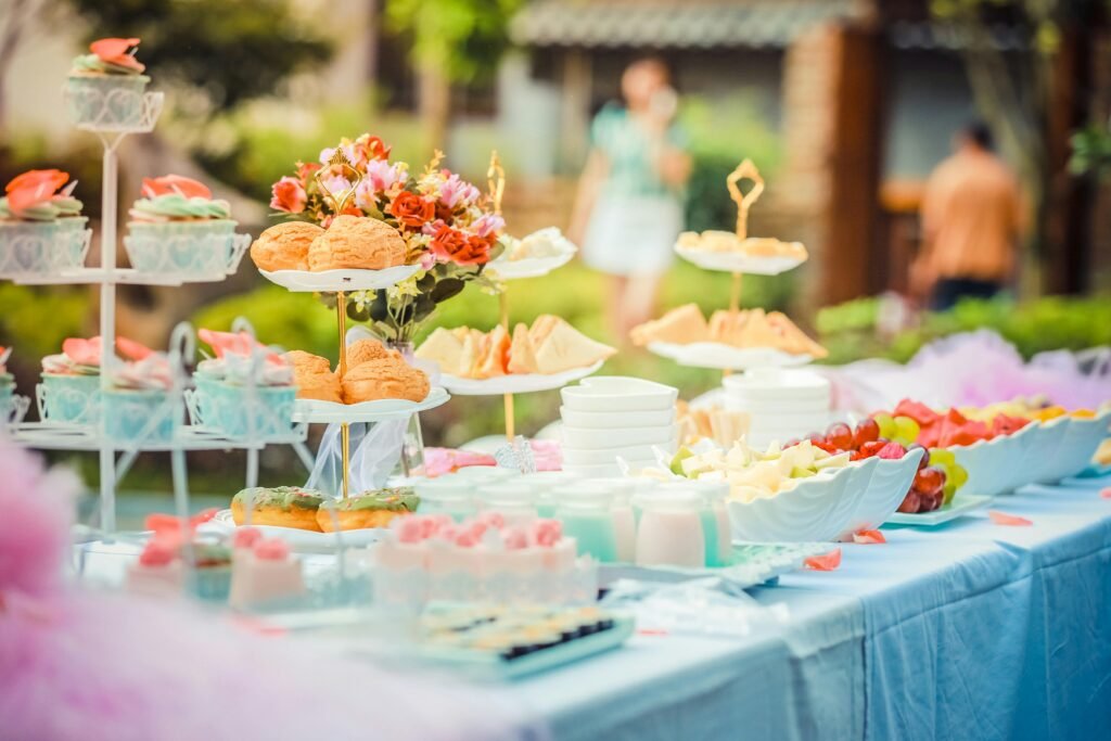 Outdoor baby shower dessert table with a variety of treats, including cupcakes, sandwiches, and fruit platters, decorated with floral arrangements.